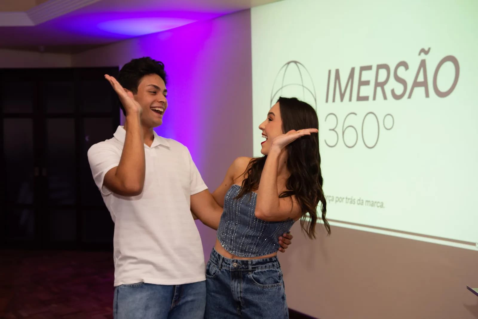 Os dois apresentadores da Liberty rindo e comemorando ao lado do telão, no fim do treinamento