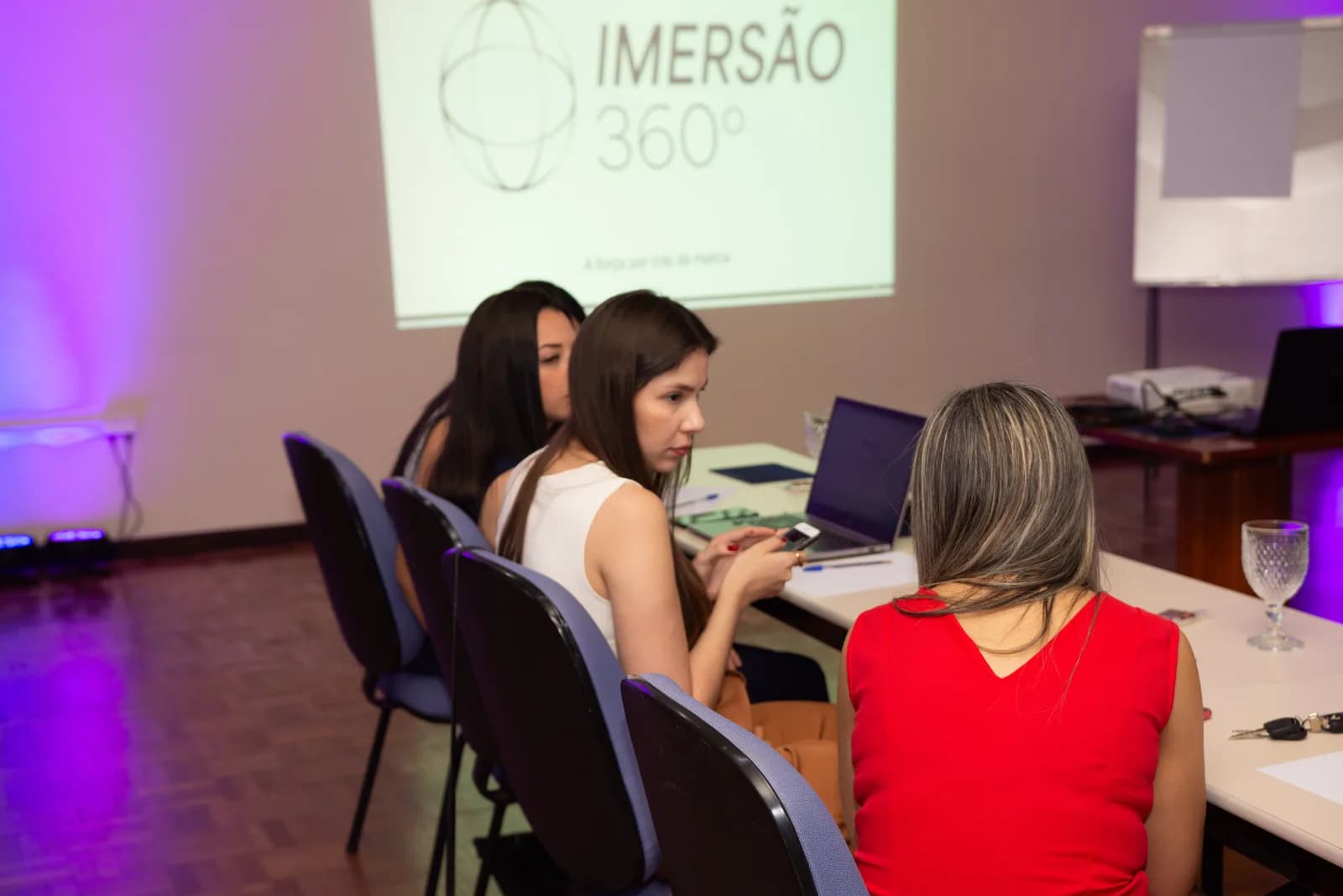 Participantes sentadas às mesas durante o treinamento, no salão de luz baixa