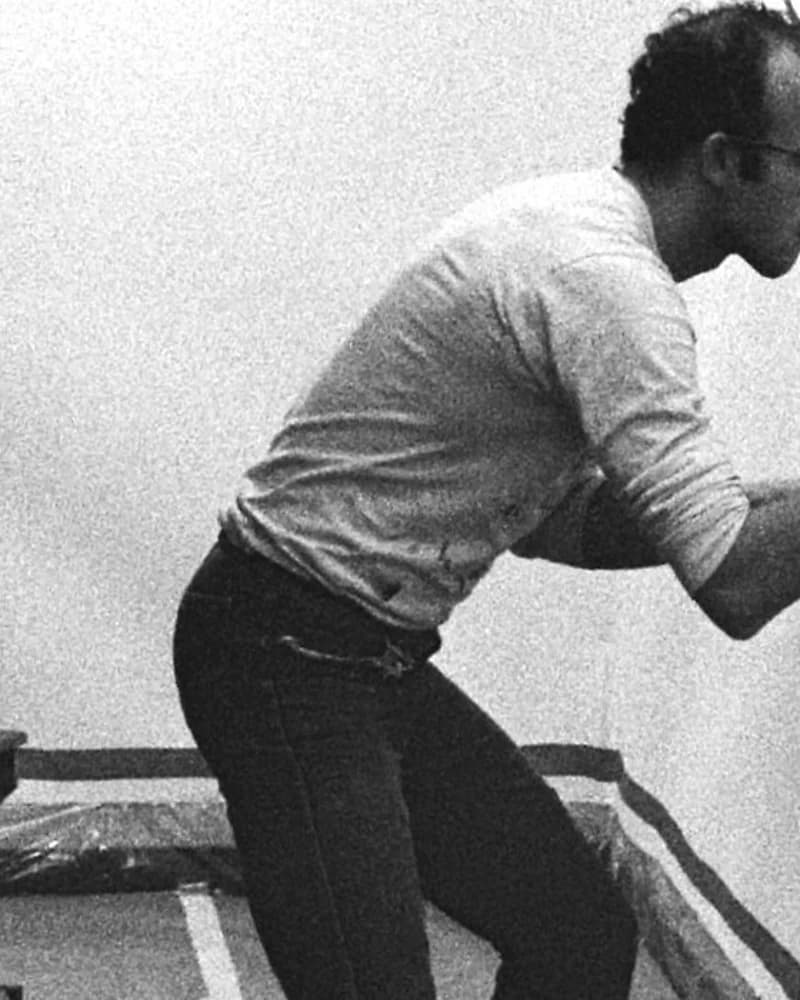 Keith Haring de perfil, curvado sobre uma parede branca com um pincel na mão, em 1986